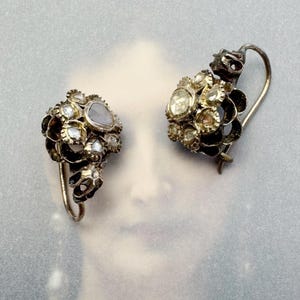 Peut inclure: Une paire de boucles d'oreilles anciennes en argent avec un groupe de petites pierres précieuses facettées. Les boucles d'oreilles ont un design floral délicat et sont accentuées par une pierre précieuse plus grande en forme de poire.