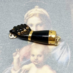 May include: A black and gold charm in the shape of a hand with a gold ring at the wrist. The charm is made of black resin and has gold accents.