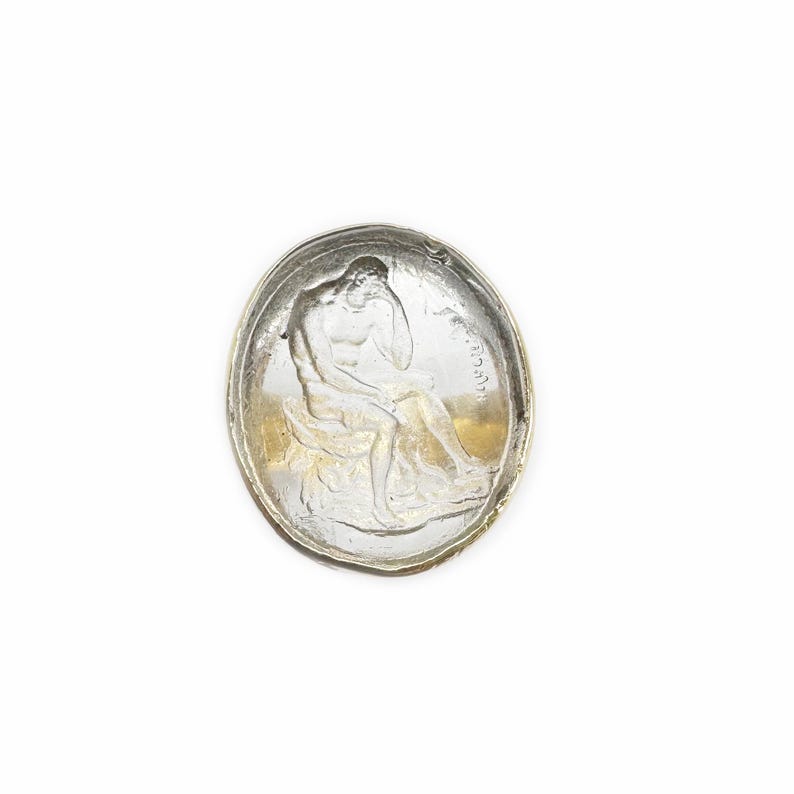 May include: A silver-toned glass paperweight featuring a raised image of a seated figure. The paperweight has a smooth, rounded surface.