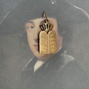 Pode incluir: Pingente dourado com os Dez Mandamentos, com algarismos romanos. O pingente é encimado por uma coroa e tem um pequeno laço para uma corrente. O fundo é um retrato de um homem.