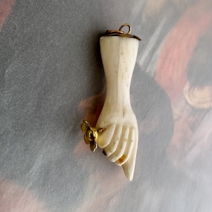May include: A carved off-white hand pendant featuring a gold-toned metal ring with a small flower design on the finger. The pendant has a gold-toned metal loop at the top for attachment, showcasing a detailed wrist and fingers in a downward position.