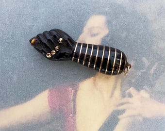 以下が含まれることがあります： 握りこぶしの形をしたダークブラウンの彫刻入りホーンペンダント。手首の部分には複数の細い水平なゴールドトーンの金属バンドが装飾されており、指の関節には小さな丸いゴールドトーンの金属スタッドがあしらわれています。