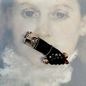 Pode incluir: Broche vintage em forma de mão preta, com detalhes prateados e duas esferas douradas. A mão é adornada com fio de prata e uma pulseira prateada. O fundo apresenta um retrato desfocado.