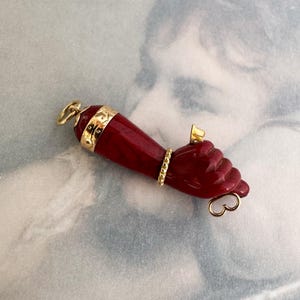 Pode incluir: Um pingente de pedra esculpida em vermelho escuro com formato de mão e antebraço. A peça apresenta detalhes em metal dourado, incluindo um punho decorativo no topo, um anel texturizado no dedo, um pequeno elemento dourado na articulação e um aro.
