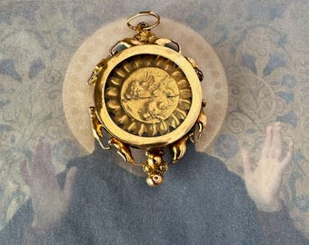 Peut inclure: Pendentif doré avec un motif circulaire. Il présente un motif de soleil et une image centrale. Il possède des détails complexes et une petite boucle pour l'accrocher. Le fond est d'une couleur douce et sourde.