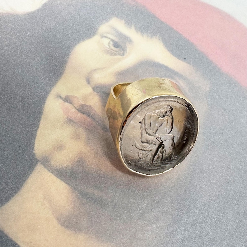 May include: A gold ring with a clear glass top featuring a detailed engraved image of a person sitting on a rock. The ring is on a background of a painting of a person's face.