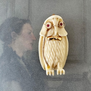 May include: Cream-coloured owl figurine with red eye accents, gold-coloured wire-rimmed spectacles, and a gold bow tie. A gold chain hangs from the spectacles. The owl is carved with a textured pattern. The background is a blurred image of a person.