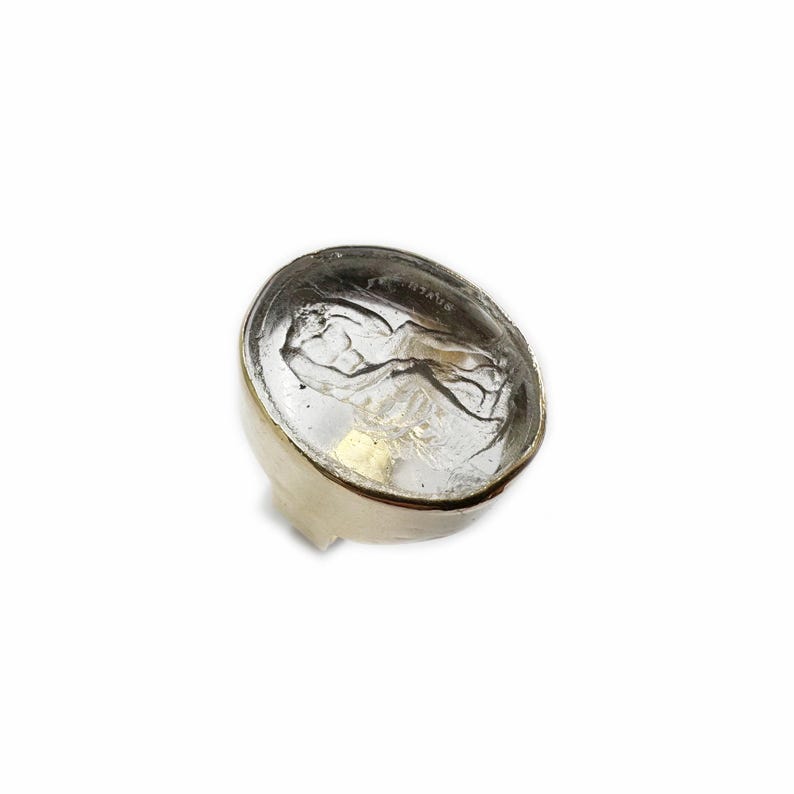 May include: A clear glass ring with a gold setting. The glass features a raised design of a figure.