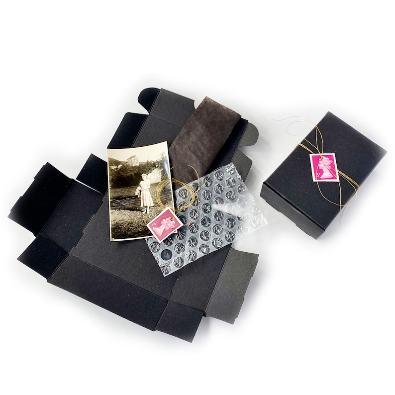 May include: A black box with a vintage photograph inside, a brown fabric piece, and bubble wrap. The box is tied with a gold ribbon and a pink stamp.
