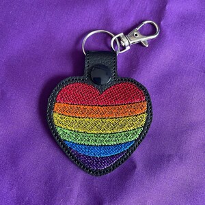 May include: A black leather heart-shaped keychain with a rainbow embroidered design. The keychain has a silver metal ring and a lobster clasp.