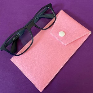 Puede incluir: Un estuche para gafas de cuero rosa con cierre de botón blanco y un par de gafas Ray-Ban negras en la parte superior.