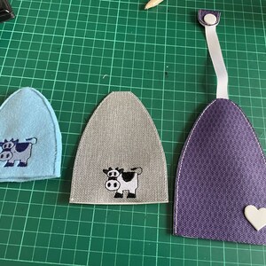 May include: Three fabric cow-shaped ornaments. One is light blue, one is gray, and one is purple with a white heart.