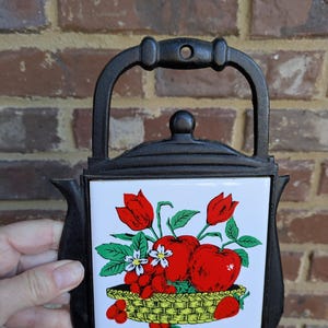 May include: Black cast iron trivet with a white tile insert featuring a colorful fruit and flower design. The tile shows red apples, tulips, and berries in a yellow basket. The trivet has a handle for easy use.