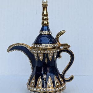 May include: A small, blue and gold decorative teapot with a pearl-like knob on the lid. The teapot is decorated with gold trim and rhinestones.