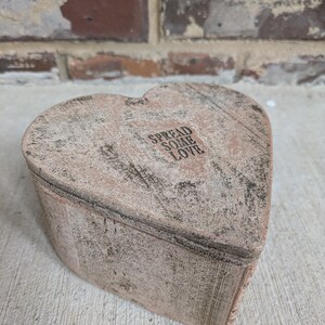 May include: A heart-shaped, weathered, light brown box with a lid. The lid has the words "SPREAD SOME LOVE" printed on it in a dark font. The box has a rustic, aged appearance.