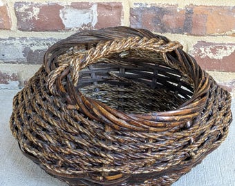May include: A woven basket with a round shape and a handle. The basket is made of brown and tan natural materials, with a textured surface. The handle is made of twisted rope. The basket is sitting on a concrete surface.