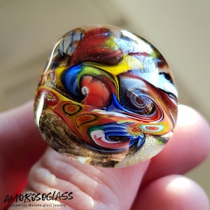 Peut inclure: Un presse-papier en verre de Murano fait à la main, aux couleurs vives. Il présente des motifs tourbillonnants de rouge, de bleu, de jaune et d'or. Le verre est tenu par un doigt, mettant en valeur les détails. Le texte "AMOROSOGLASS" est visible.