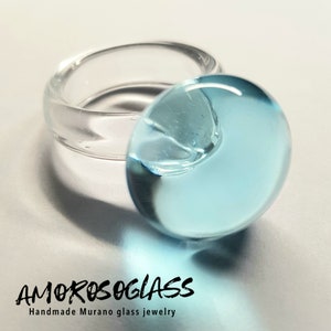 Op de afbeelding: Een heldere glazen ring met een lichtblauwe, afgeronde middenstuk. De ring is handgemaakt en voorzien van de tekst "AMOROSOGLASS" en "Handmade Murano glass jewelry". Een uniek sieraad.