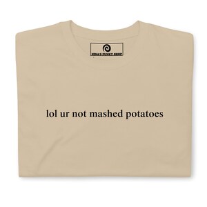 Può includere: Una maglietta beige con la scritta "lol ur not mashed potatoes" stampata sul davanti. La maglietta ha un piccolo logo in alto che dice "Nina's Funky Shop".
