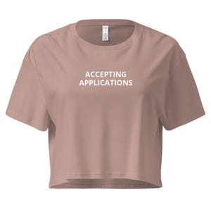 May include: A dusty rose crop top with short sleeves. The shirt features the text "ACCEPTING APPLICATIONS" in white block letters across the front. The shirt has a crew neck and a relaxed fit.