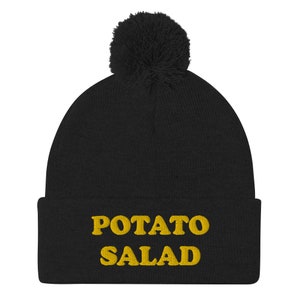 Puede incluir: Gorro de punto negro con un pompón esponjoso en la parte superior. El gorro tiene las palabras "POTATO SALAD" bordadas en amarillo en la parte delantera.