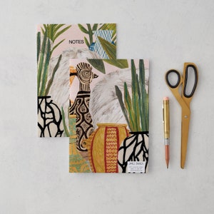 May include: Two colorful notebooks with a botanical illustration of plants and a bird in a pot. The notebook on top has the word "NOTES" printed on the cover. The notebook on the bottom has the text "JAMES BARKER" printed on the cover.
