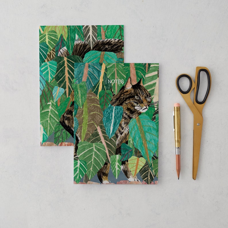 May include: Two green and brown illustrated notebooks with a cat and foliage design. The top notebook has the word "NOTES" printed on it.