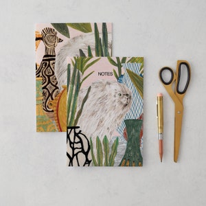 May include: Two illustrated notebooks with a pink and white background. The top notebook features a bird with a long neck and a cat with a white and gray fur. The bottom notebook features a cat with white and gray fur, plants, and the word "NOTES".