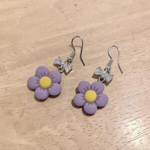 Op de afbeelding: Een paar hangende oorbellen met een paarse bloemhanger met vijf bloemblaadjes en een geel hartje. Elke oorbel heeft een zilverkleurige metalen haak en een kleine, heldere, gefacetteerde kristallen strik boven de bloemvorm.
