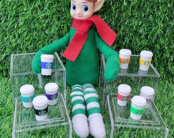 May include: A green elf doll with a red scarf is sitting on a table with multiple miniature coffee cups. The cups are on clear acrylic shelves. The cups have a green and white logo.
