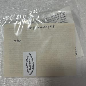 May include: A clear plastic bag containing several pieces of paper with Hebrew text. The main piece of paper is off-white with black text. A small white sticker with black text is affixed to the paper. The text is blurry.