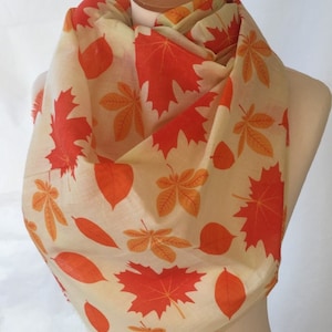 May include: A cream-coloured scarf featuring a repeating pattern of orange and red autumn leaves. The scarf is draped over a mannequin, highlighting the fabric's design and texture. The leaves are of various shapes and sizes.