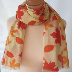 May include: A beige scarf featuring a pattern of orange and red autumn leaves. The scarf is displayed on a mannequin and has a fringed edge. The fabric is lightweight, suitable for cooler weather. The scarf measures approximately 180 cm in length.