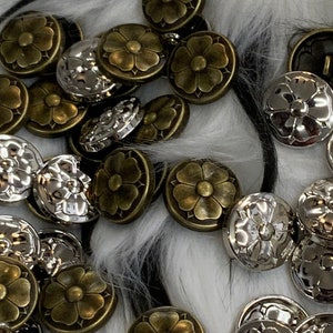 May include: A collection of silver and gold metal buttons with a flower design. The buttons are scattered on a white and black fuzzy surface.