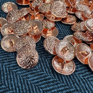 May include: A collection of rose gold metal buttons with a Celtic knot design. The buttons are round with a shank on the back for sewing. The buttons are scattered on a dark teal herringbone fabric.