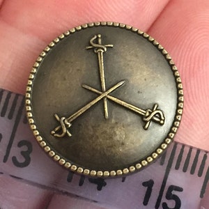 May include: A bronze-colored button with a design of three swords crossed in a triangular pattern. The button has a beaded edge.