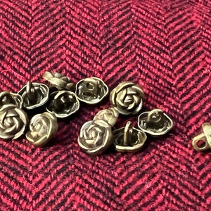 May include: A group of 10 antique gold-toned rose-shaped buttons with a decorative shank on the back. The buttons are arranged in a cluster on a red and black herringbone fabric.
