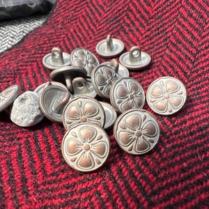 Op de afbeelding: Een verzameling metalen knopen in antieke stijl met een bloemmotief. De knopen hebben zilver- en bronskleurige tinten en liggen op een rood-zwarte visgraatstof. De knopen hebben een lus aan de achterkant om op te naaien.
