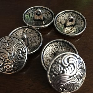 May include: A collection of six silver-colored metal buttons. The buttons have a detailed floral and swirl design on the front. The backs of the buttons have a textured appearance with a loop for sewing.