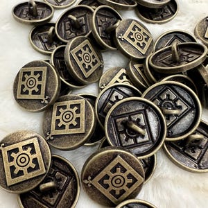 Puede incluir: Un conjunto de botones de color latón antiguo. Cada botón presenta un diseño geométrico en relieve dentro de un cuadrado y un ojal en la parte posterior para coser. Los botones tienen un aspecto vintage y envejecido.