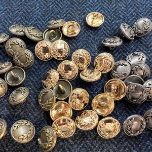 May include: A collection of gold and silver metal buttons with a laurel wreath design. The buttons are round and have a raised design.