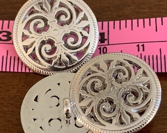 May include: Three silver-colored, ornate buttons with a filigree design. The buttons are round with a decorative border and a shank on the back. A pink measuring tape is in the background.