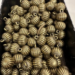 Puede incluir: Una colección de pequeñas cuentas en forma de calabaza, de color latón antiguo. Cada cuenta tiene estrías verticales y un lazo en la parte superior para ensartar. Las cuentas están contenidas en una caja negra, lo que sugiere que son para manualidades o joyería.