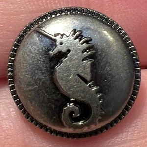 May include: A silver-toned button with a raised design of a seahorse. The button has a decorative border.