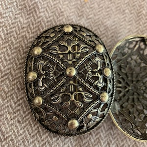 May include: A close-up of a decorative, oval-shaped, metal brooch with intricate, Celtic-inspired designs. The brooch is made of a brass-coloured metal and features a circular pattern with a central knot.