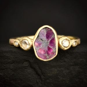 Könnte beinhalten: Ein goldener Ring mit einem großen, unregelmäßig geformten, rosa-violetten Edelstein in der Mitte. Der Ring wird von drei kleineren, klaren Edelsteinen auf jeder Seite flankiert. Der Ring ist vor einem schwarzen Hintergrund platziert.