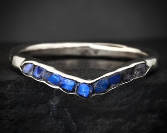 Puede incluir: Un anillo de plata en forma de chevrón con una hilera de pequeñas piedras azules de corte rugoso incrustadas a lo largo del centro en forma de V. La banda de metal tiene un acabado pulido y las piedras muestran un color azul intenso.