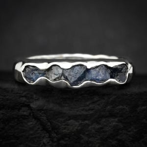 May include: A silver ring with a row of dark blue, irregularly shaped stones. The ring has a smooth, polished band and a textured setting for the stones. The stones have a rough, natural appearance, contrasting with the polished metal.