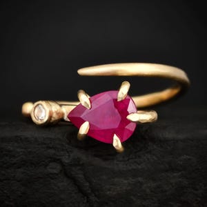 Op de afbeelding: Een gouden bypass-ring met een peervormige, dieproze robijn die door vier klauwen wordt vastgehouden. De ringband heeft een getextureerde, matte afwerking en bevat een kleine, ronde, heldere edelsteen die in de zijkant van de ring is gezet.