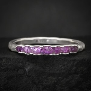 May include: A silver ring with a line of vivid, rough-cut purple gemstones. The band has a textured, organic look, and the gemstones are set close together. The ring is shown against a dark backdrop, emphasising its colour and design.
